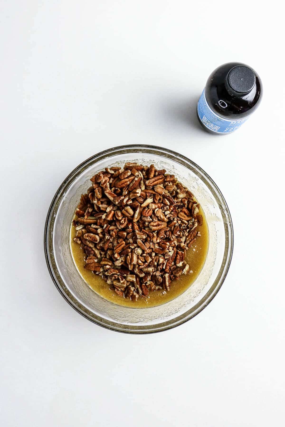 A bowl of pecans in liquid next to a bottle of maple syrup on a white background.