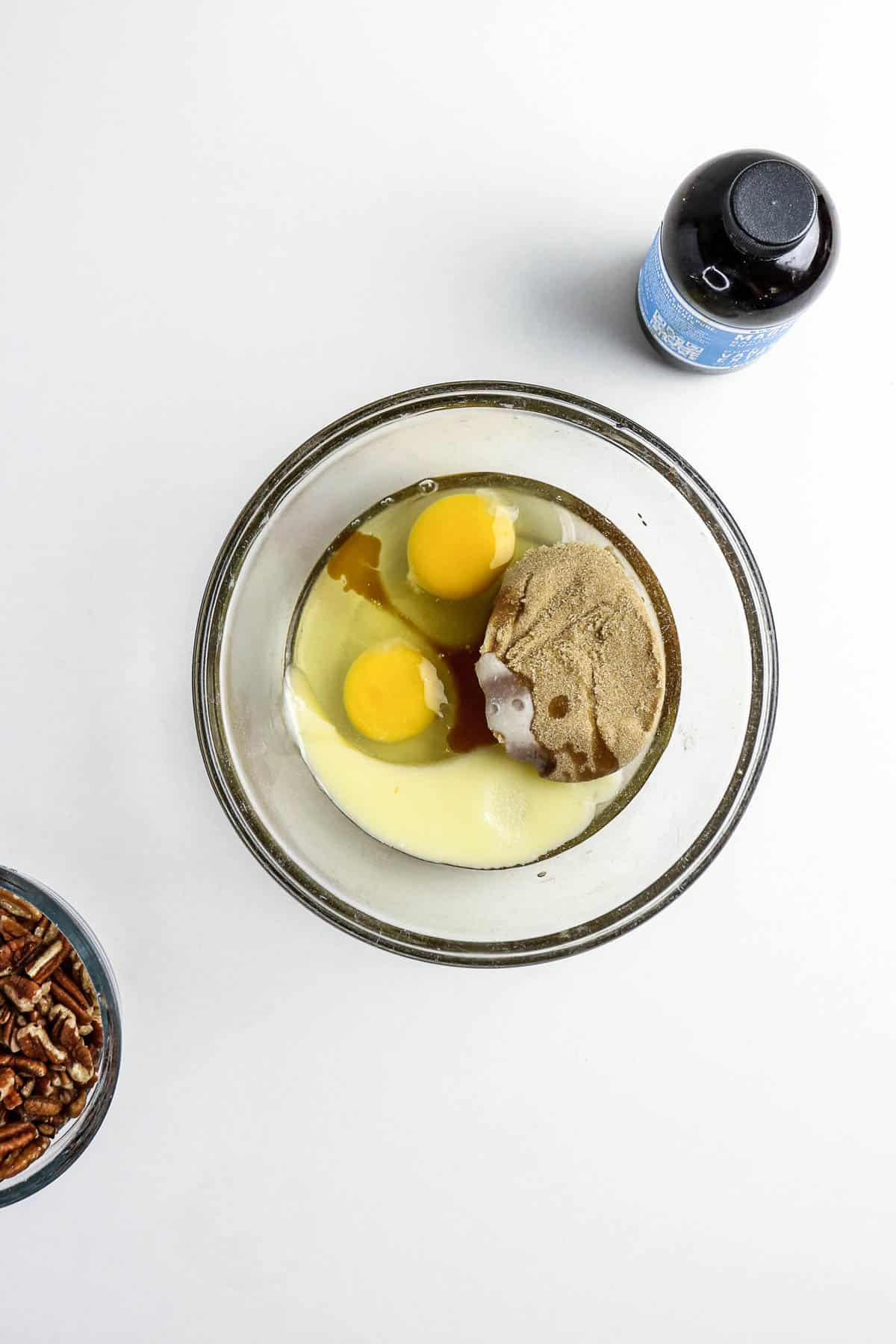 Glass bowl with eggs, brown sugar, and liquid ingredients. Bottle and pecans are nearby on a white surface.