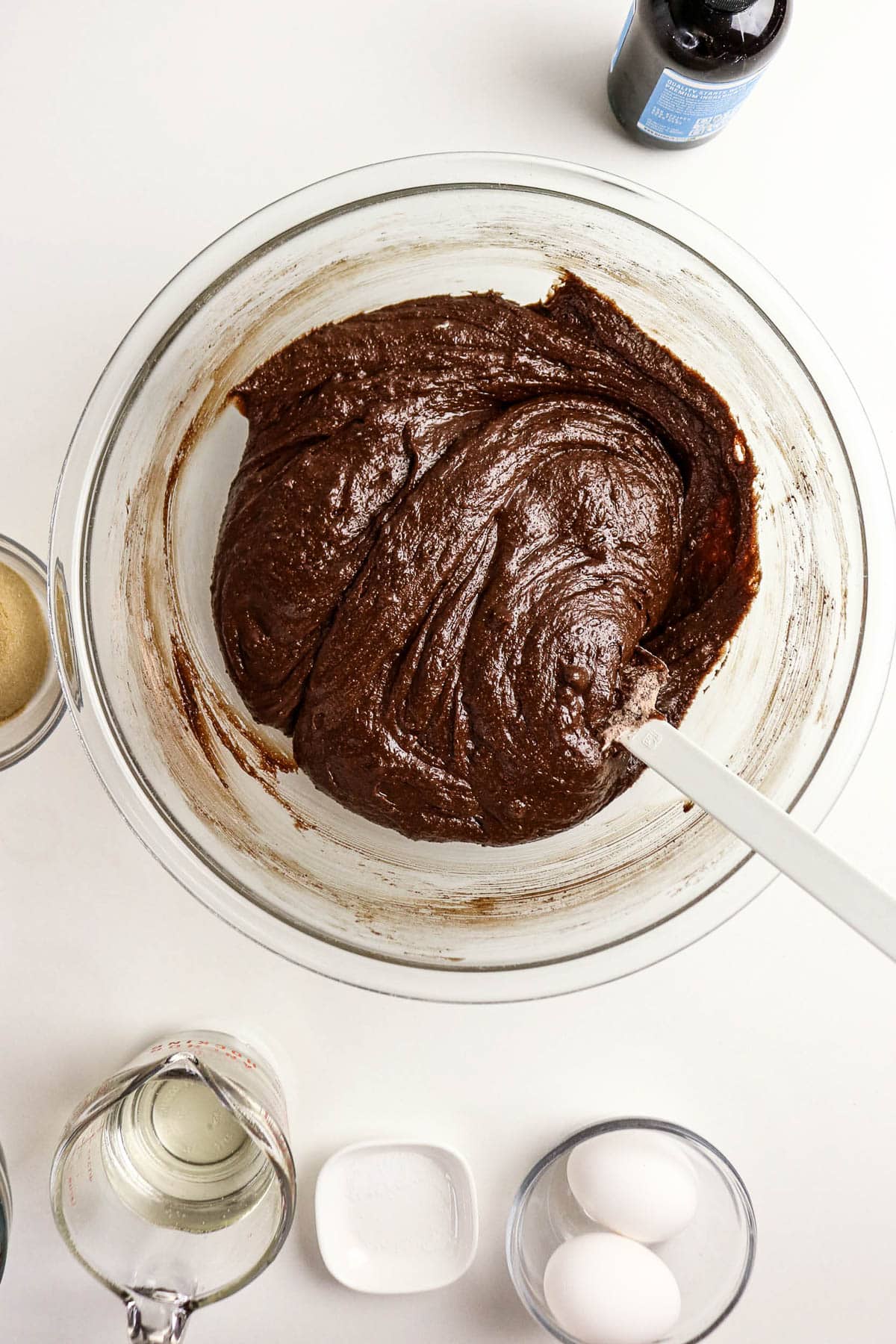 A glass bowl of brownie batter with a spatula, surrounded by eggs, sugar, oil, and vanilla on a white surface.