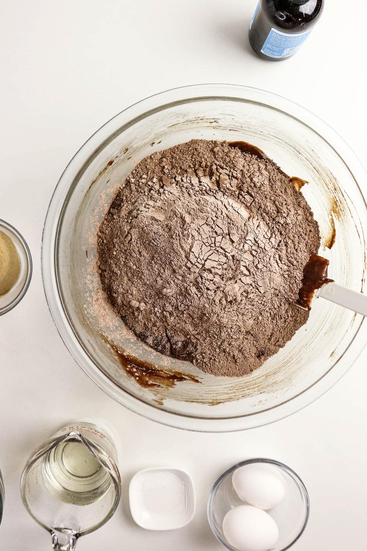 A mixing bowl with dry cocoa ingredients, surrounded by eggs, sugar, oil, vanilla, and a measuring cup.