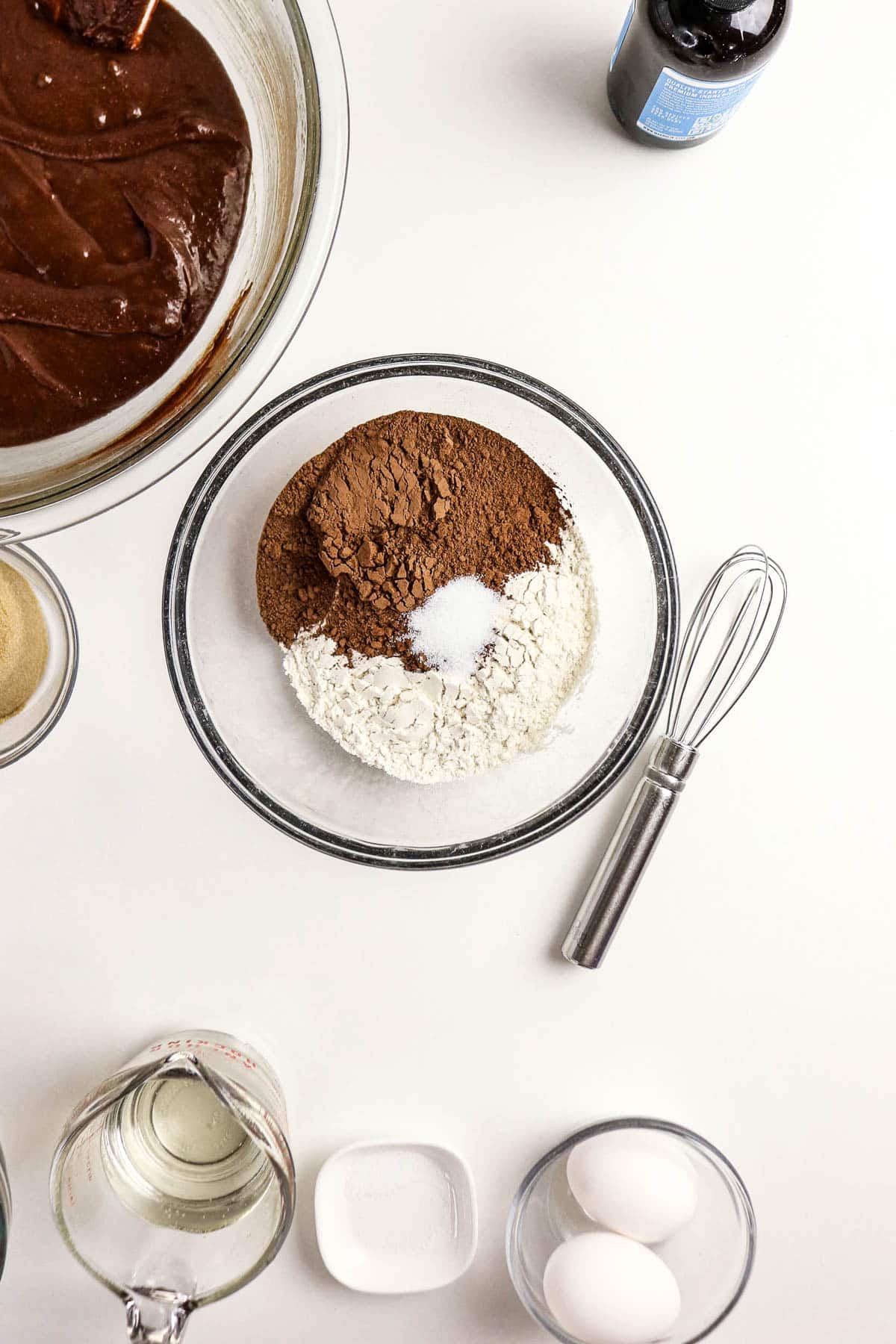 Bowl with cocoa powder, flour, and salt next to a whisk, surrounded by baking ingredients on a white surface-perfect for whipping up delicious Pecan Pie Brownies.