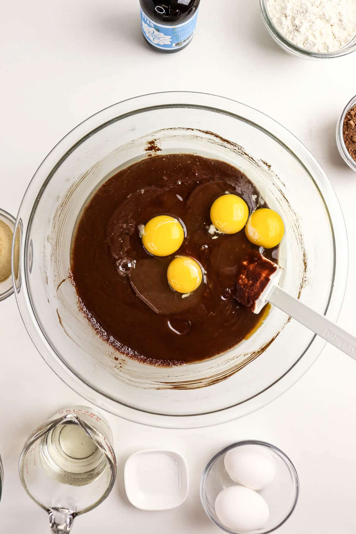 A mixing bowl with chocolate batter and four eggs, surrounded by baking ingredients on a white surface.