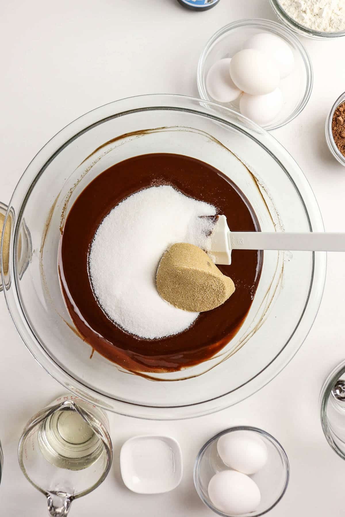 A bowl of chocolate batter with sugar being mixed in, surrounded by eggs, flour, and other baking ingredients.