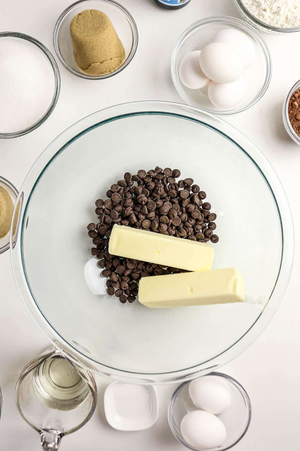 A glass bowl with butter and chocolate chips surrounded by eggs, sugar, flour, and other baking ingredients.