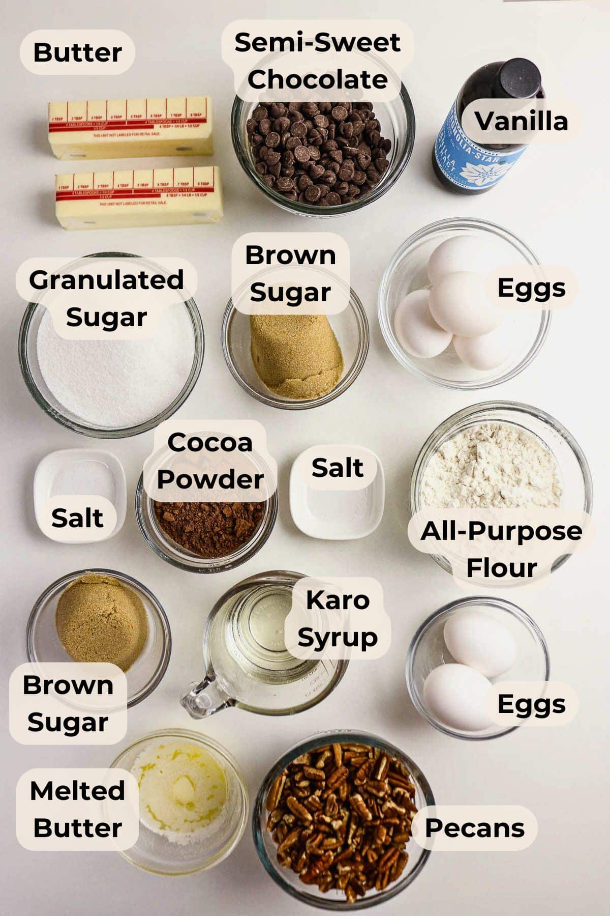 Overhead view of labeled baking ingredients in bowls and jars, including butter, chocolate, sugar, eggs, and pecans.