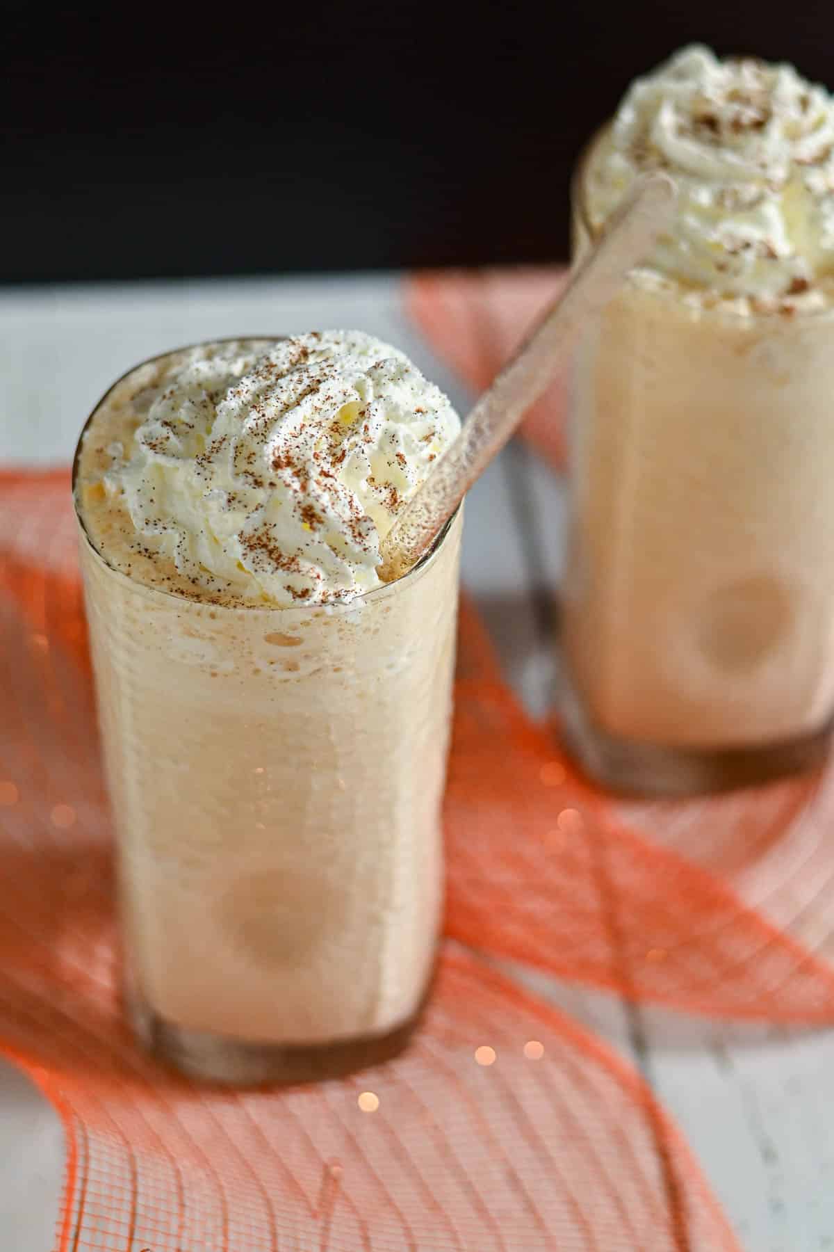 Two tall glasses of creamy milkshake topped with whipped cream and cinnamon, served with straws on an orange mat.