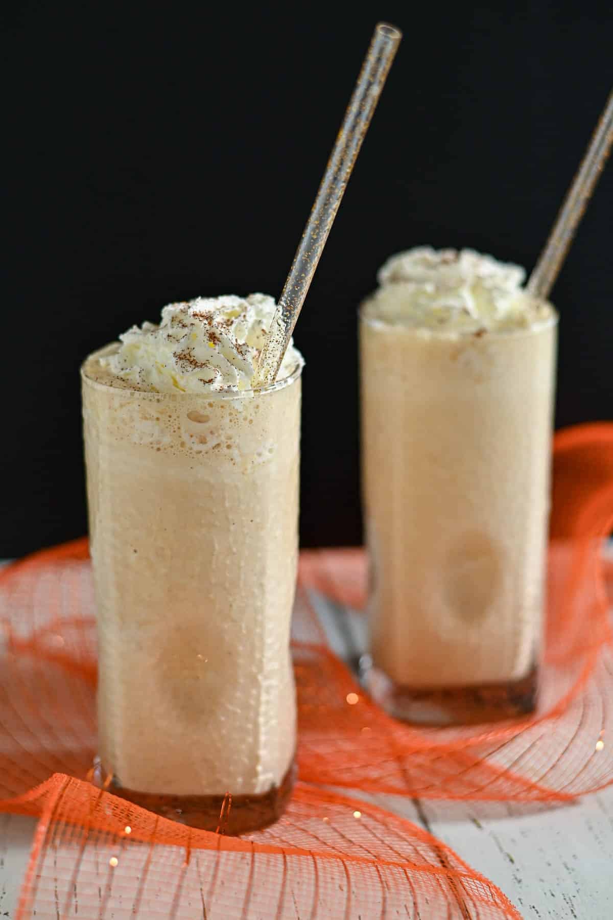 Two tall glasses of creamy pumpkin milkshake topped with whipped cream and straws, set on a white table with orange decor.