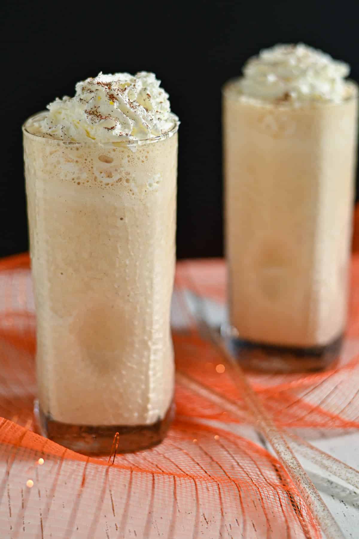 Two tall glasses of creamy iced coffee topped with whipped cream, set on a table with orange netting decor.