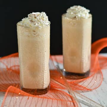 Two tall glasses of frothy iced coffee topped with whipped cream on a white table with orange mesh decor.