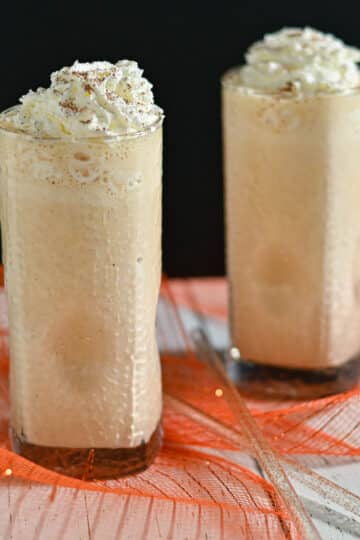 Two tall glasses of frothy iced coffee topped with whipped cream on a white table with orange mesh decor.