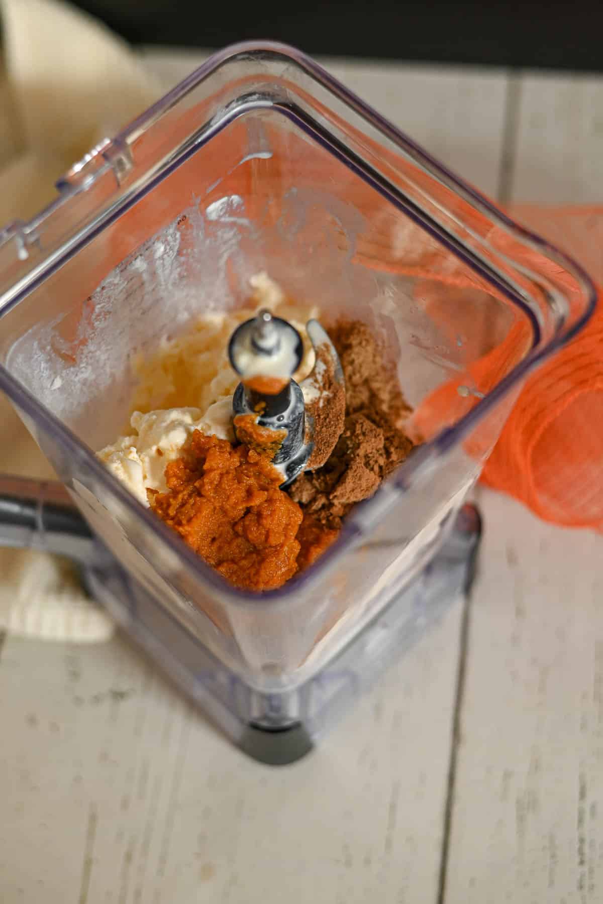 A blender filled with cream, pumpkin puree, and spices on a white wooden surface.