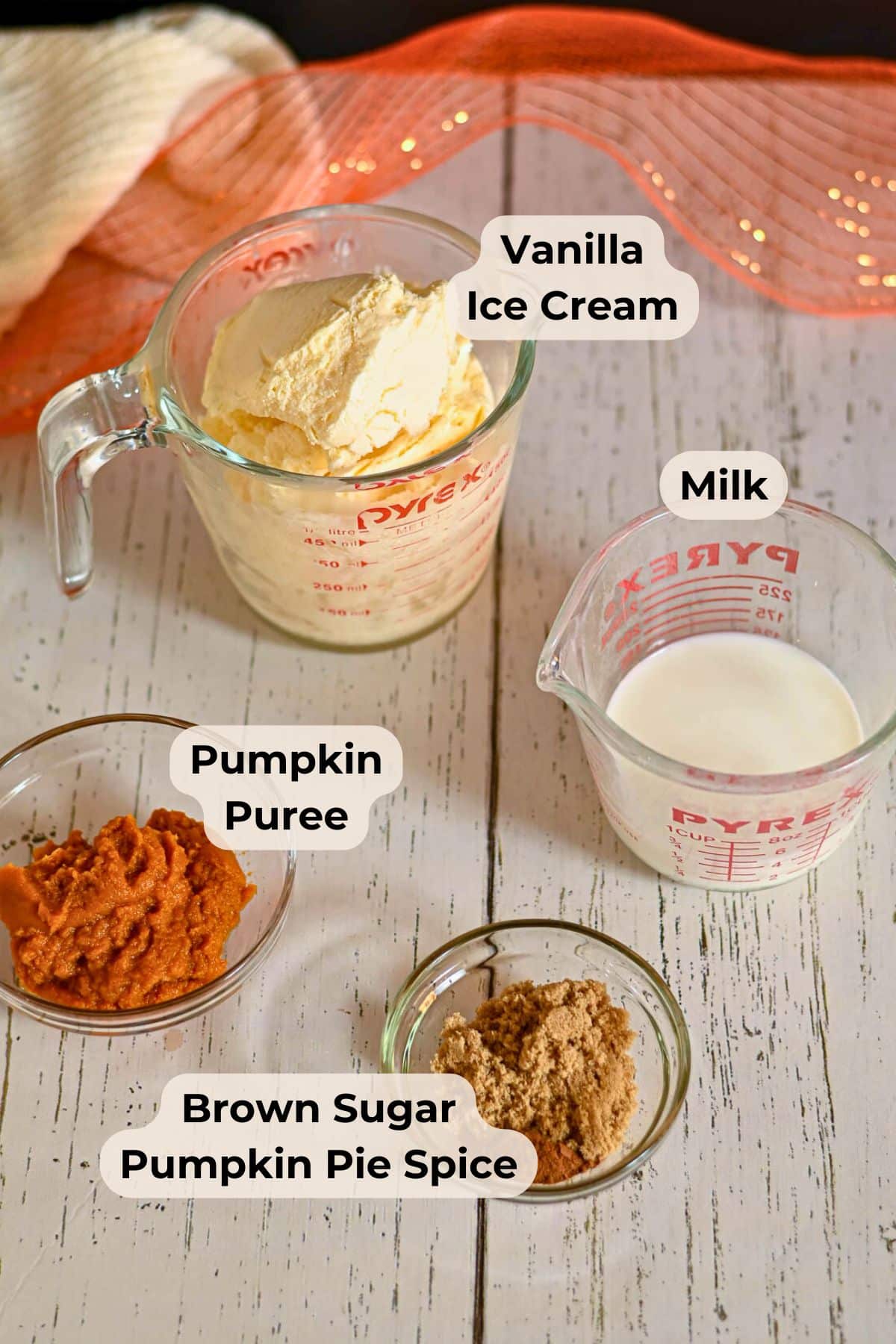 Measuring cups and bowls with vanilla ice cream, milk, pumpkin puree, brown sugar, and pumpkin pie spice on a table.