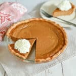 Pumpkin pie with a slice cut out, topped with whipped cream, on a glass pie plate and white napkin.