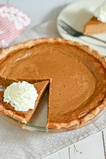 Pumpkin pie with a slice cut out, topped with whipped cream, on a glass pie plate and white napkin.