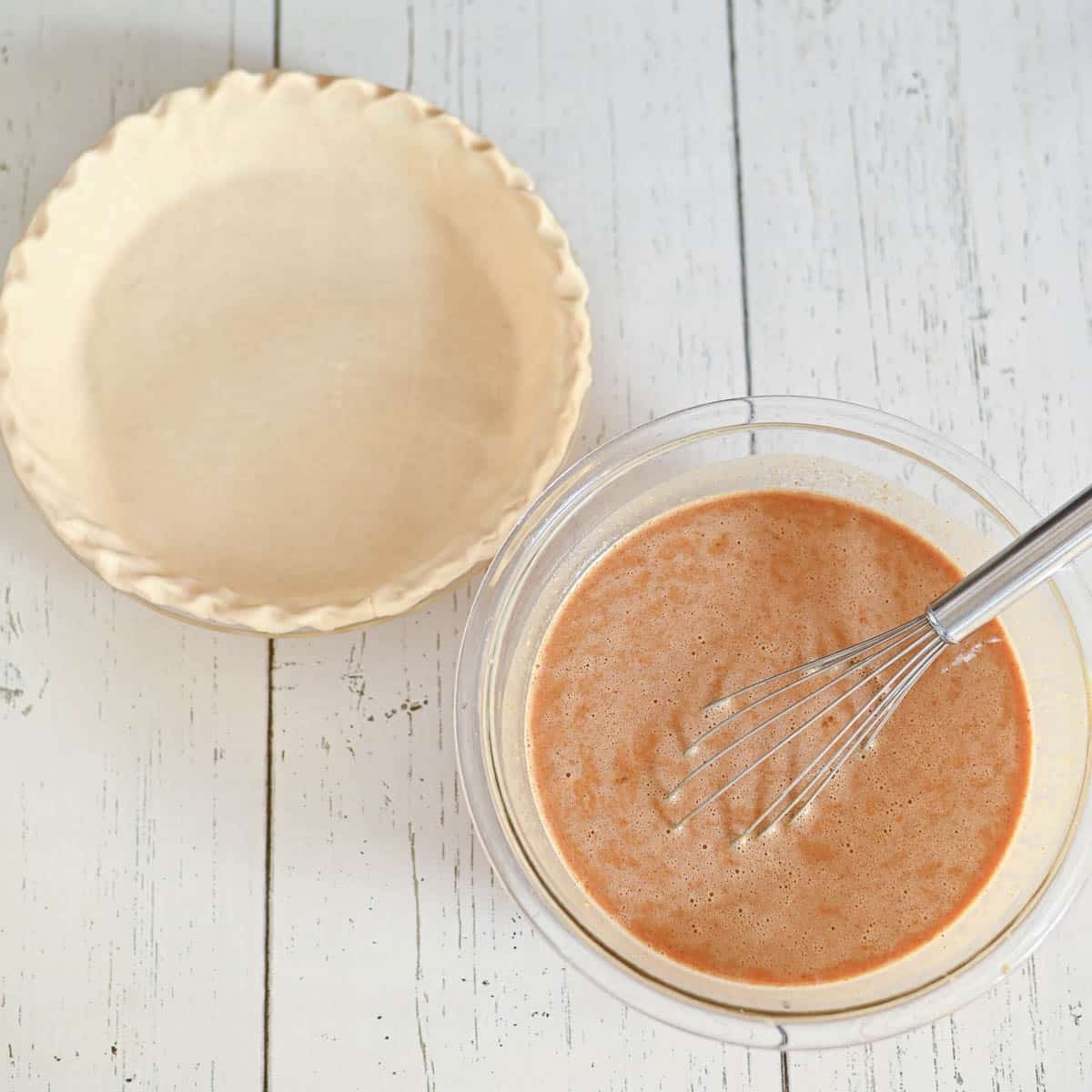 Unbaked pie crust next to a bowl of mixed pie filling with a whisk on a white wooden surface.