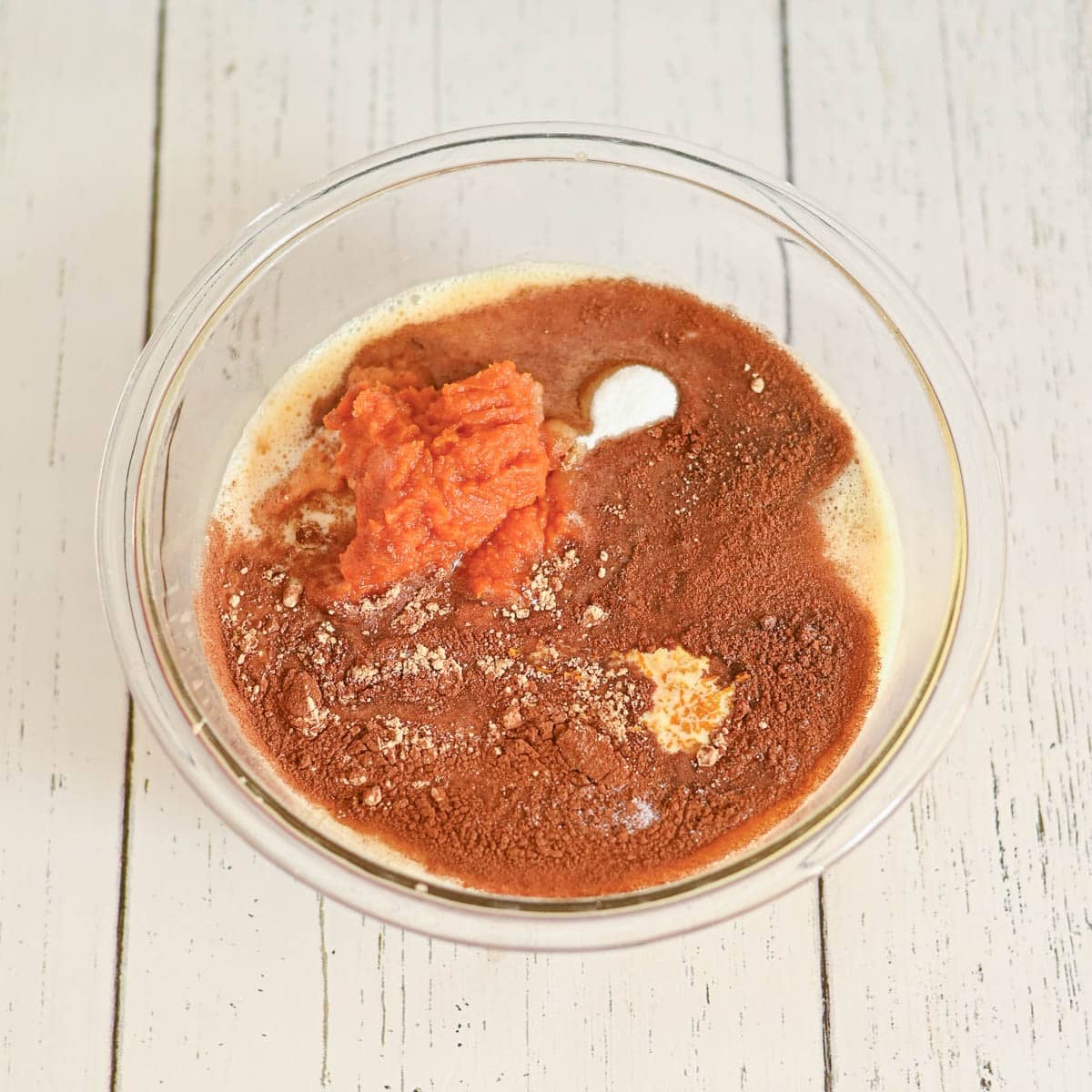 A glass bowl with pumpkin puree, cocoa powder, and other baking ingredients on a white wooden surface.