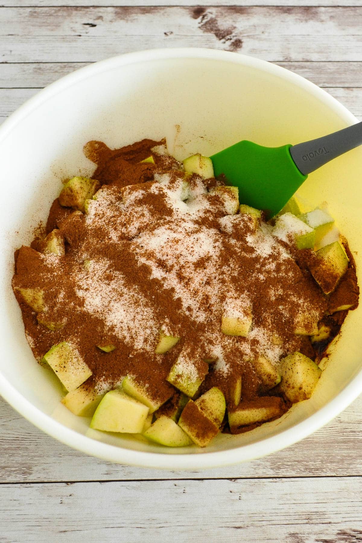 Chopped apples in a bowl topped with cinnamon and sugar, with a green spatula on the side.