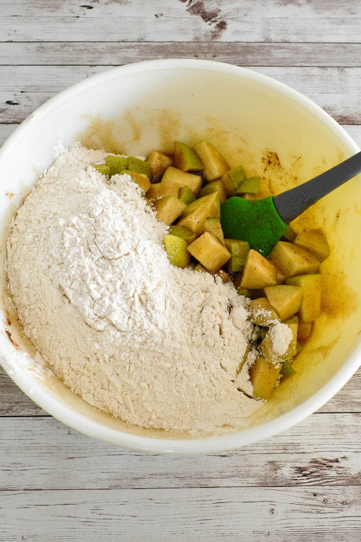 A mixing bowl with chopped apples, flour, and a spatula on a rustic wooden surface.