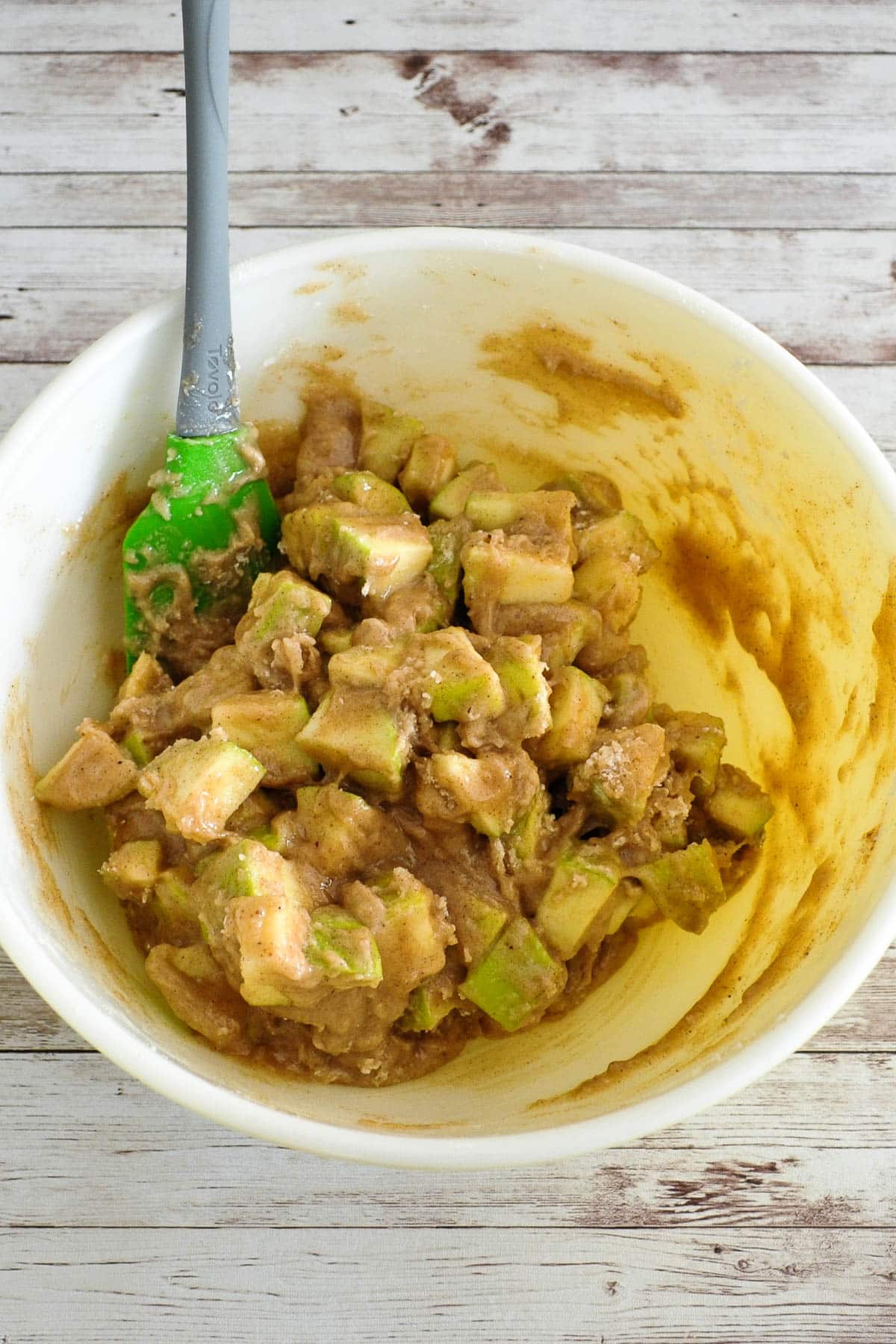 A mixing bowl with chopped green apples coated in a brown batter and a green spatula inside.
