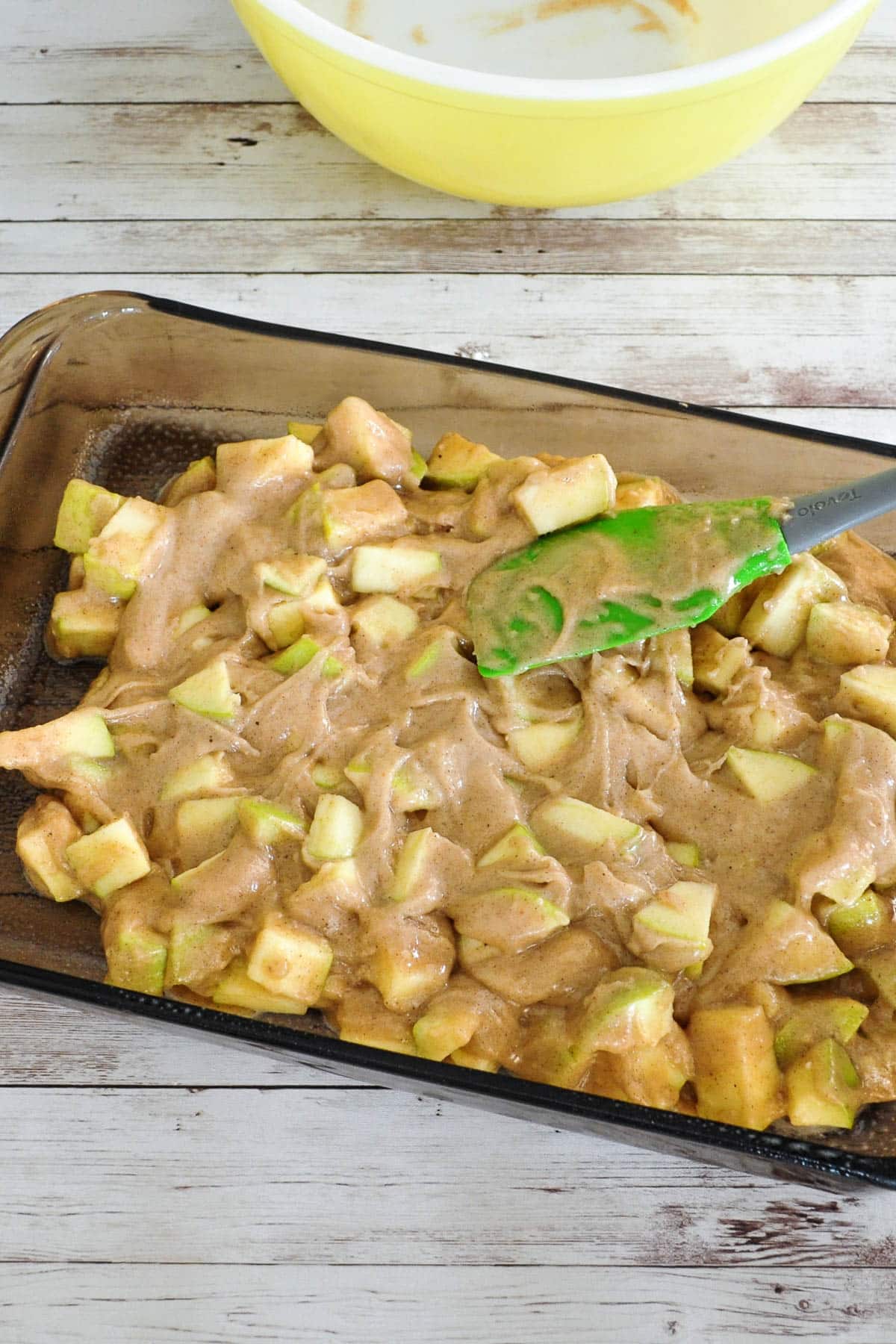 A glass baking dish filled with chopped apples and batter being spread with a green spatula.