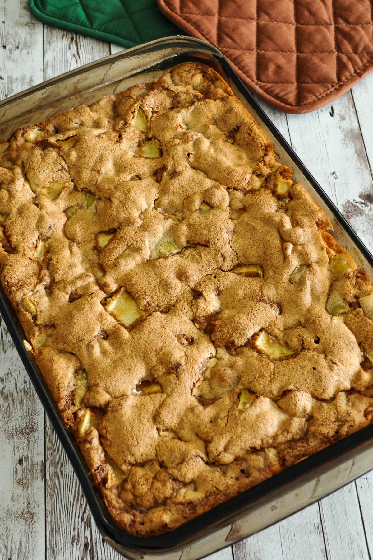 A baked apple dessert in a glass dish on a rustic wooden table with oven mitts nearby.