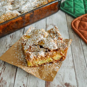 A slice of apple cake with powdered sugar on brown paper, next to a baking dish and oven mitts.
