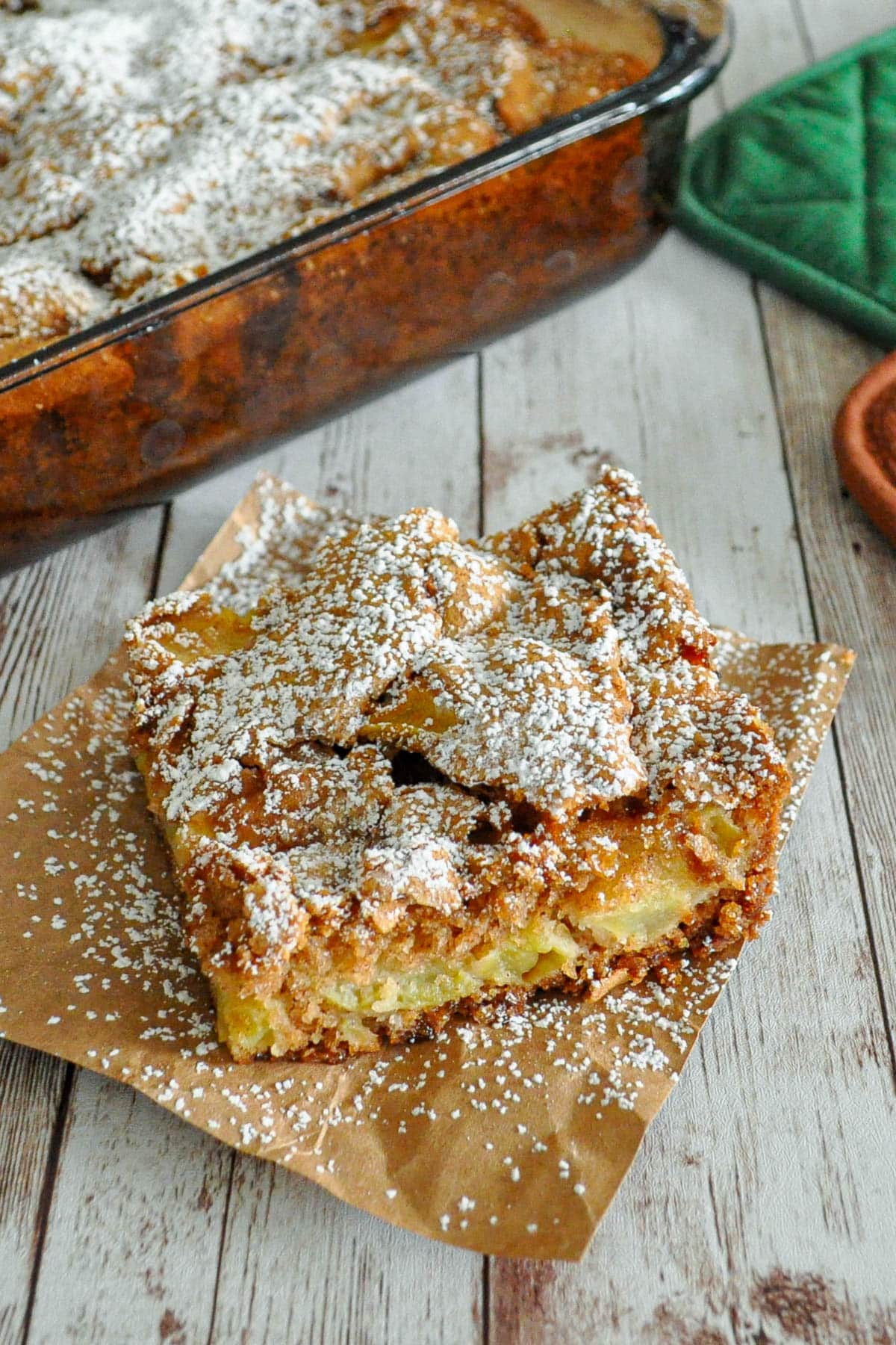 A square of apple cake topped with powdered sugar on brown paper, with a baking dish in the background.