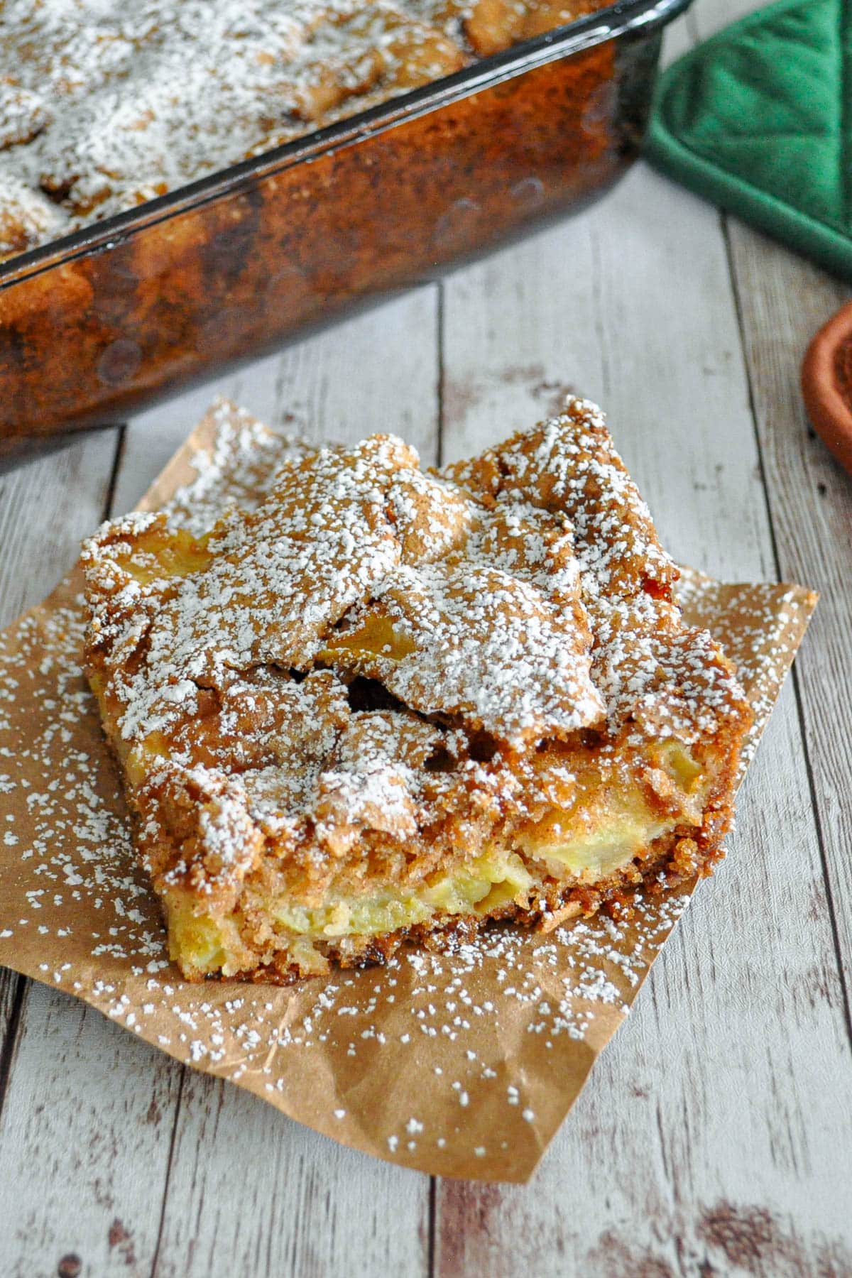 A slice of apple cake with powdered sugar on top, placed on brown parchment paper.