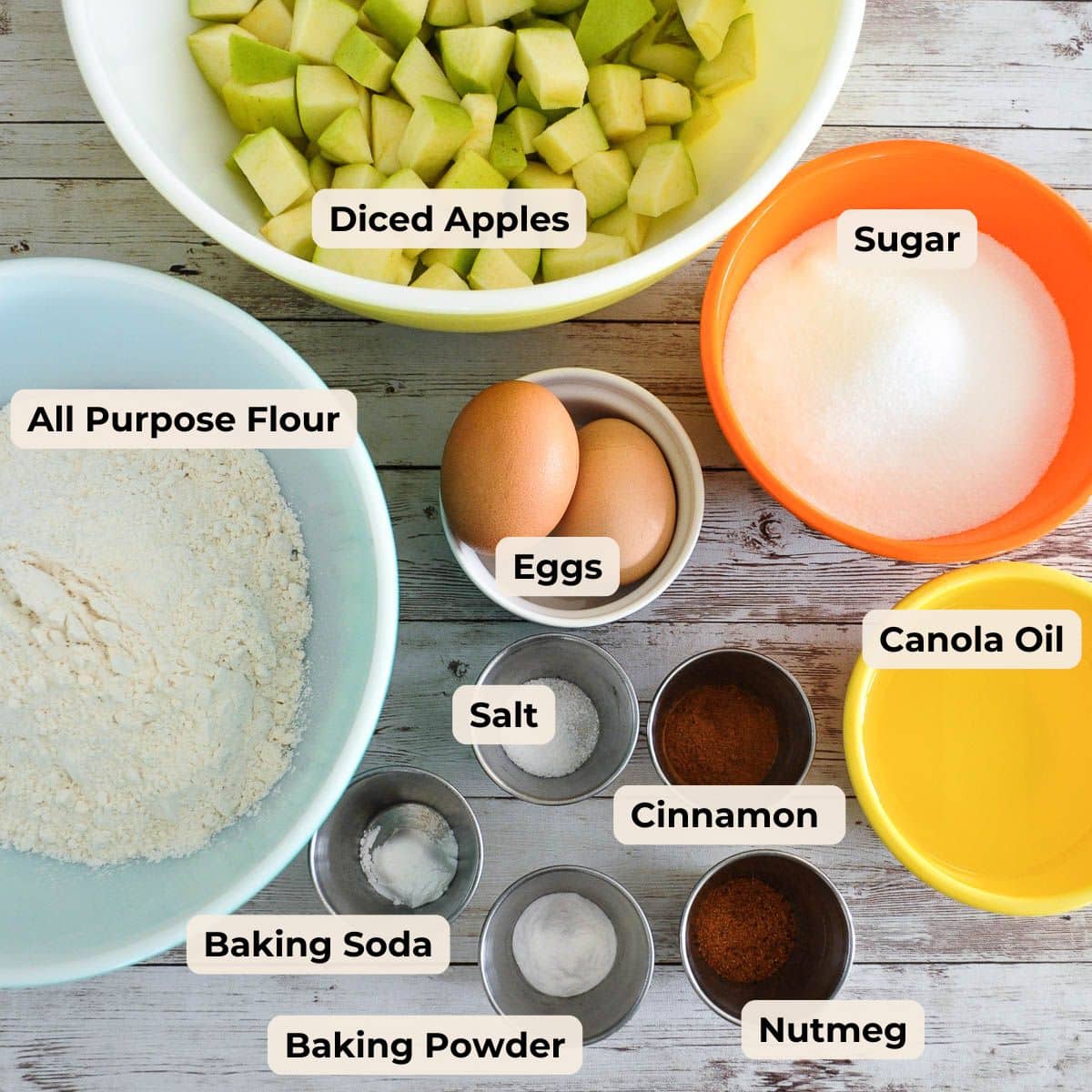 Bowls of diced apples, sugar, flour, eggs, canola oil, and small cups of spices, salt, baking soda, and baking powder.