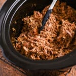 Shredded pulled pork in a slow cooker with black tongs resting on the meat.