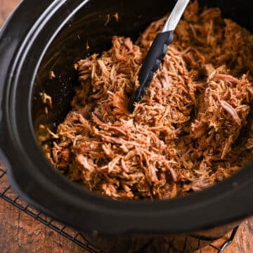 Shredded pulled pork in a slow cooker with black tongs resting on the meat.