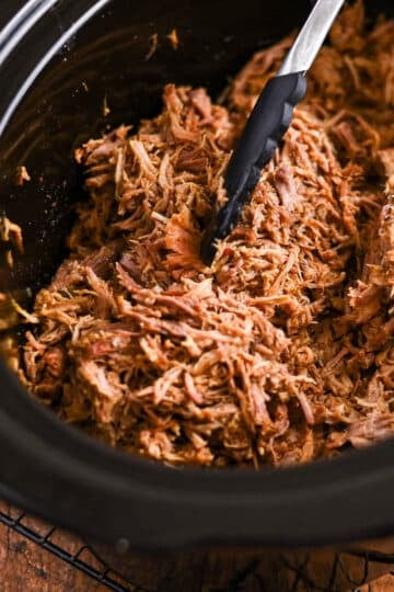 Shredded pulled pork in a slow cooker with black tongs resting on the meat.