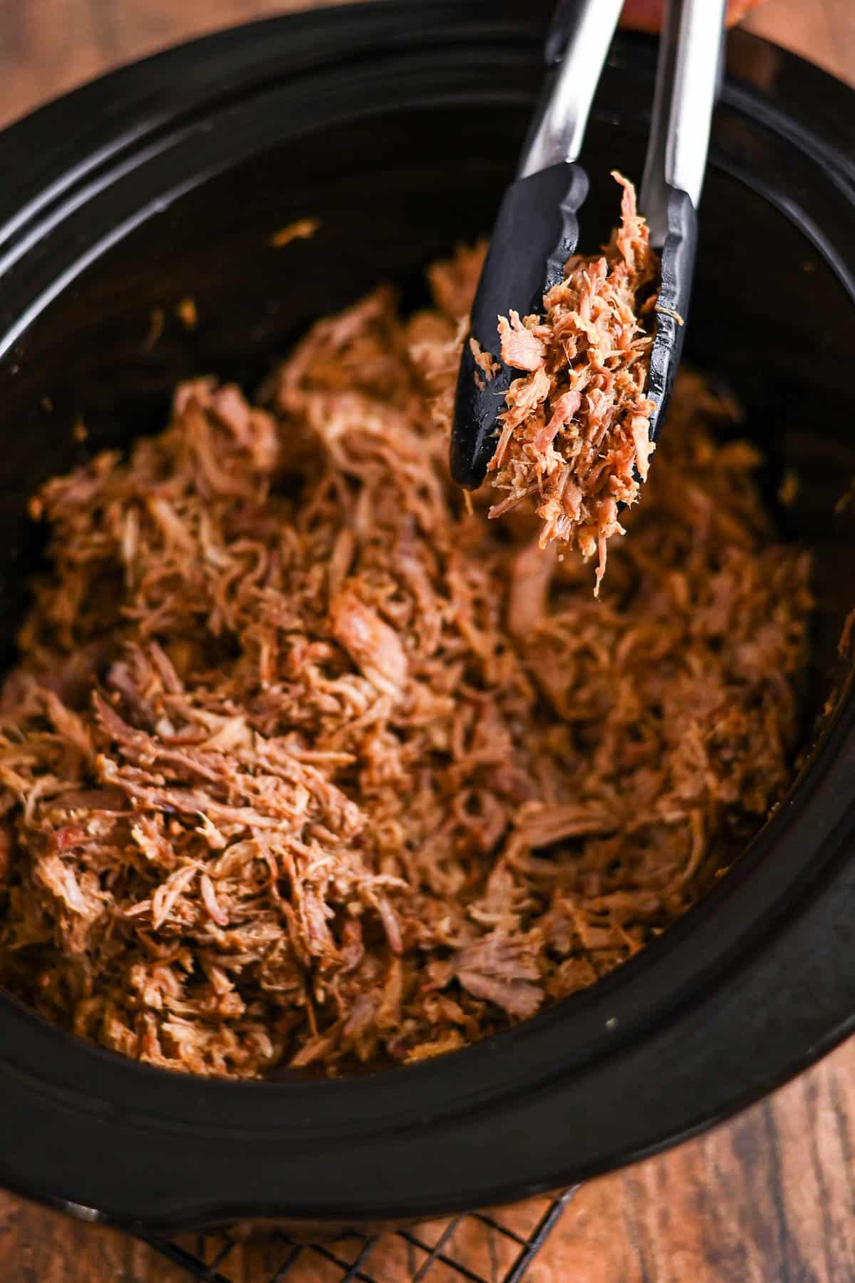 Shredded cooked meat in a slow cooker, with tongs lifting some of the meat.