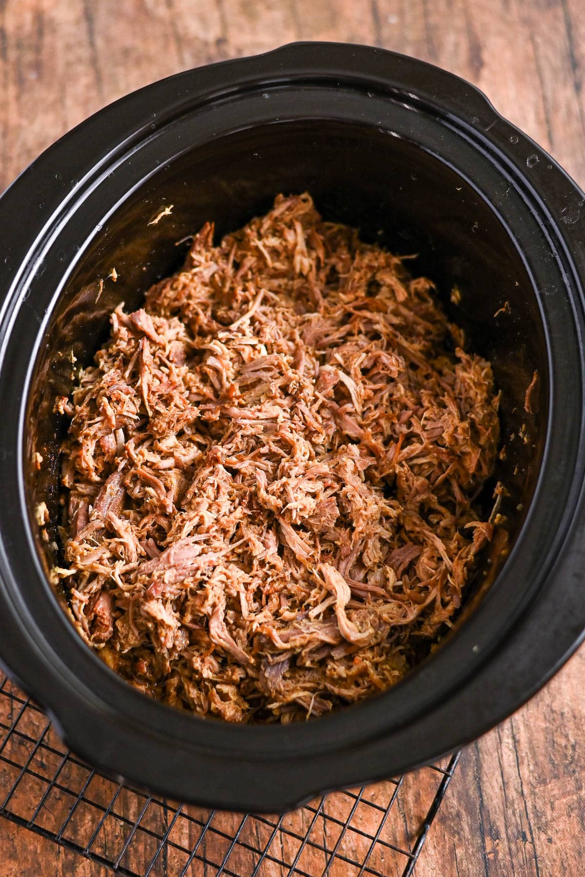 Shredded cooked beef inside a black slow cooker, placed on a rustic wooden surface.