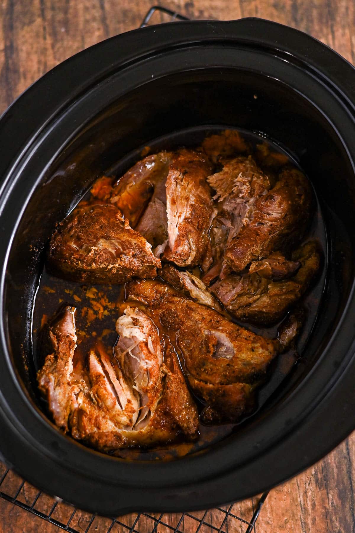 Shredded, seasoned pork cooking in a black slow cooker on a wooden surface.