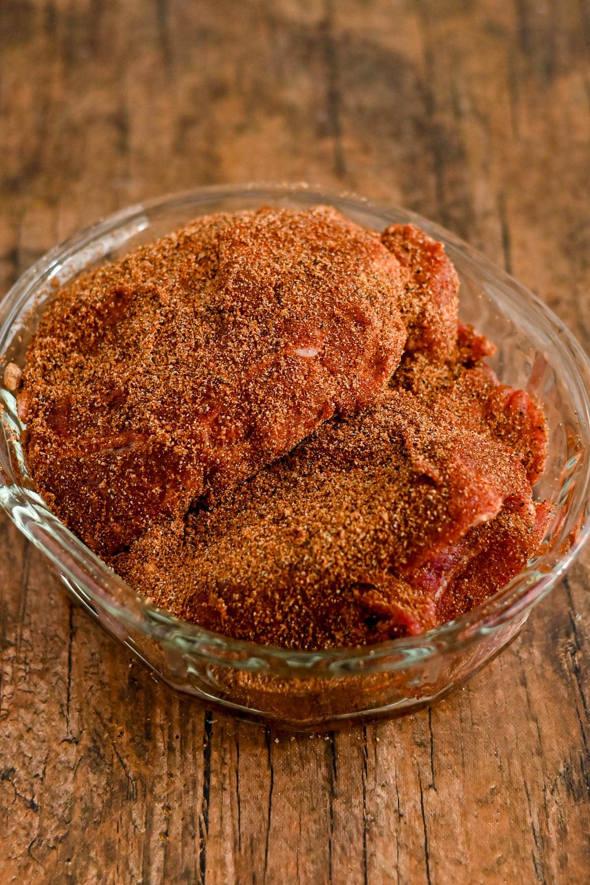Raw meat coated with dry spice rub in a clear glass bowl on a rustic wooden surface.