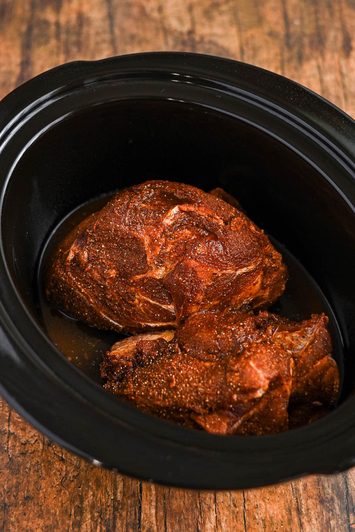 Two seasoned pieces of pork in a slow cooker with some liquid, ready to become tender and flavorful Slow Cooker Pulled Pork, on a wooden surface.