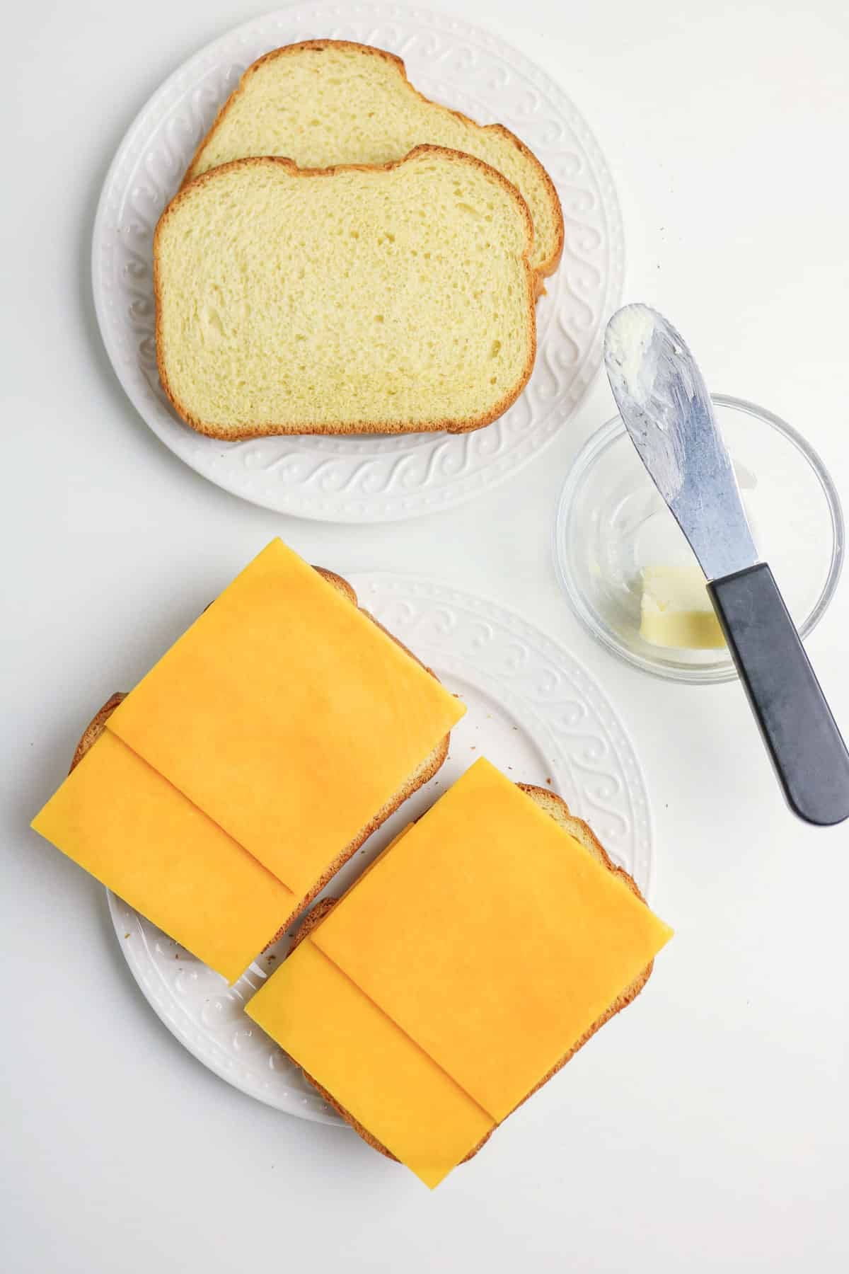Two slices of bread topped with cheddar cheese, next to plain bread and a butter knife with a small glass bowl.