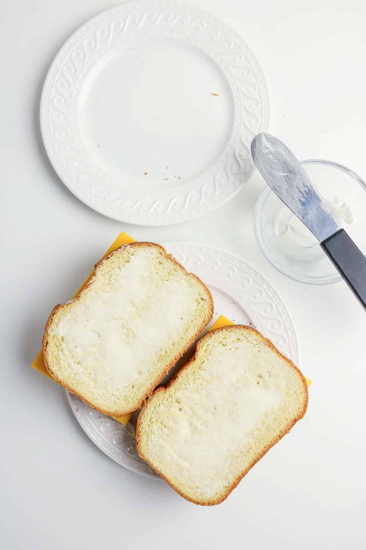 Two slices of bread with cheese and spread, next to a knife and small bowl on white plates.