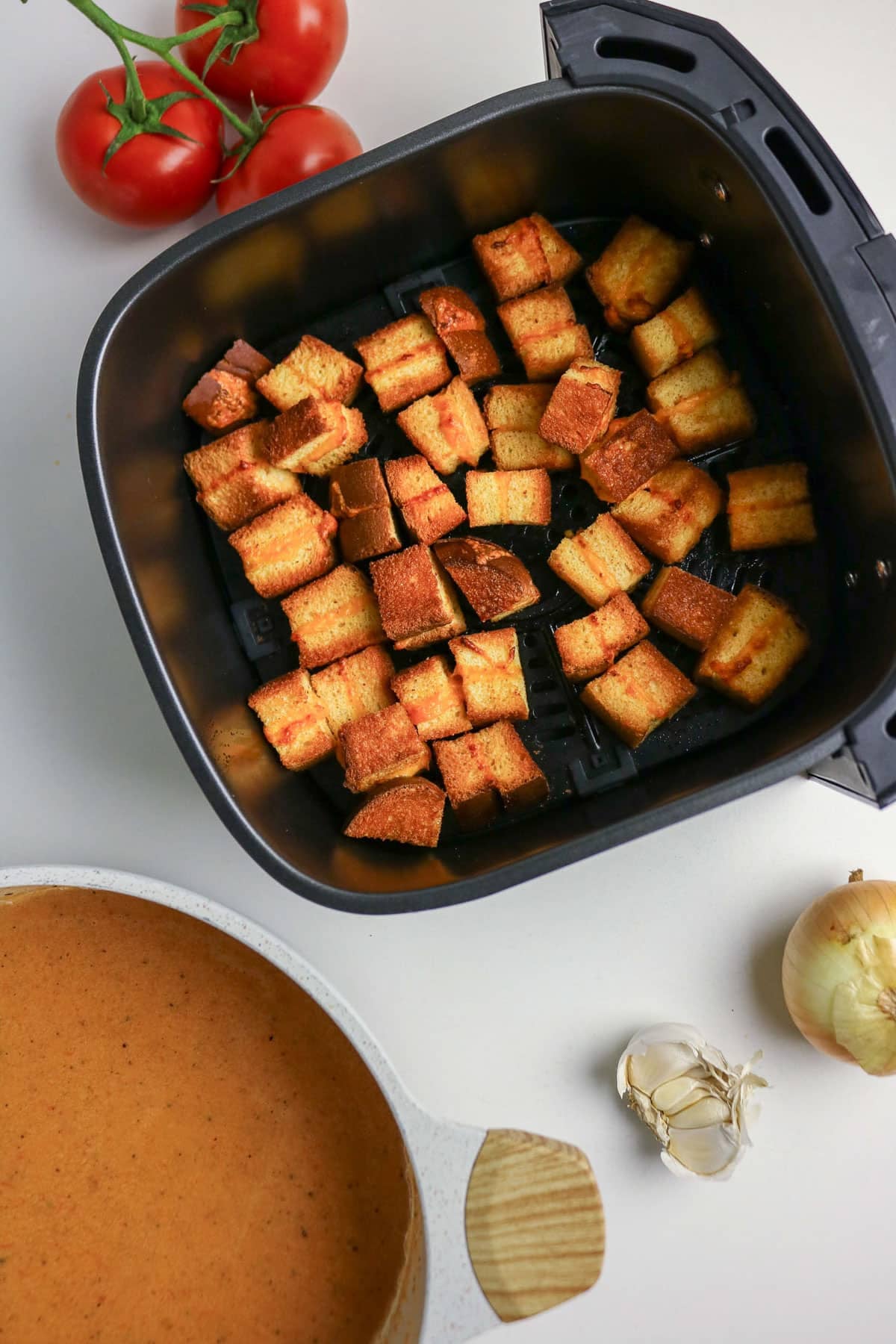 Air fryer with crispy grilled cheese croutons, fresh tomatoes, soup, onion, and garlic on a white surface.