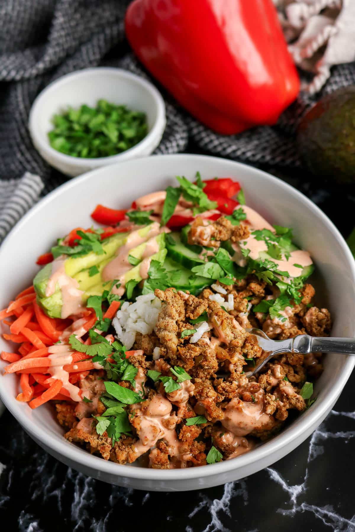 A bowl with ground meat, rice, sliced avocado, carrots, red peppers, sauce, and chopped cilantro.