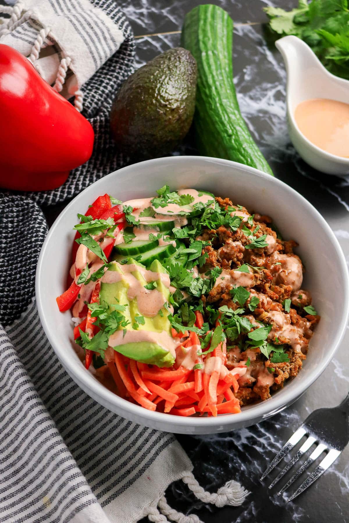 A bowl of salad with avocado, shredded carrots, red bell pepper, herbs, and creamy dressing on a marble table.
