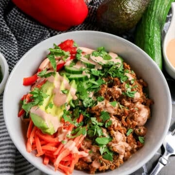 A bowl with seasoned ground meat, avocado, cucumber, red pepper, carrots, cilantro, and creamy dressing.