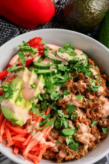 A bowl with seasoned ground meat, avocado, cucumber, red pepper, carrots, cilantro, and creamy dressing.