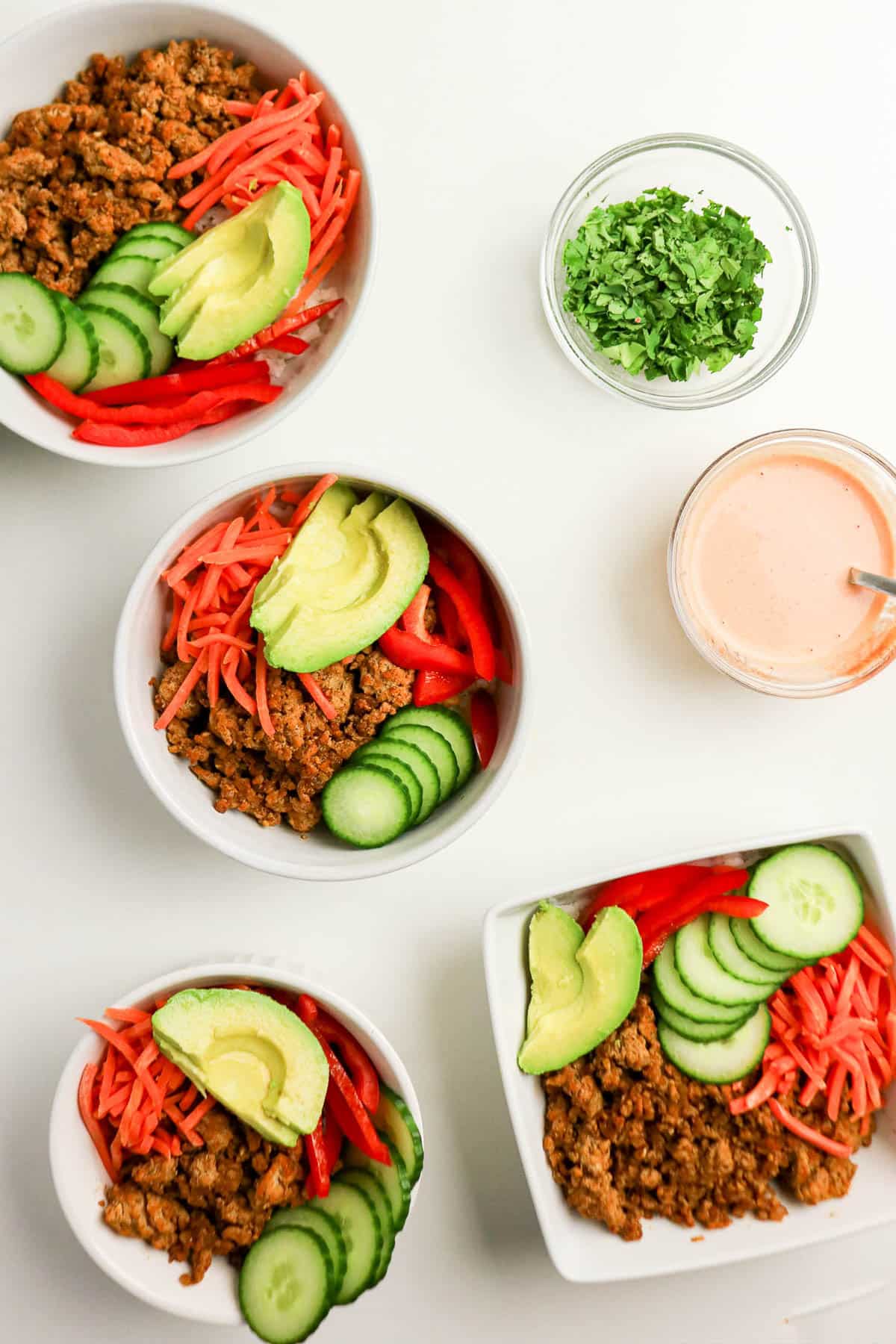 Four bowls with taco meat, sliced avocado, cucumber, and red peppers, plus cilantro and sauce on the side.