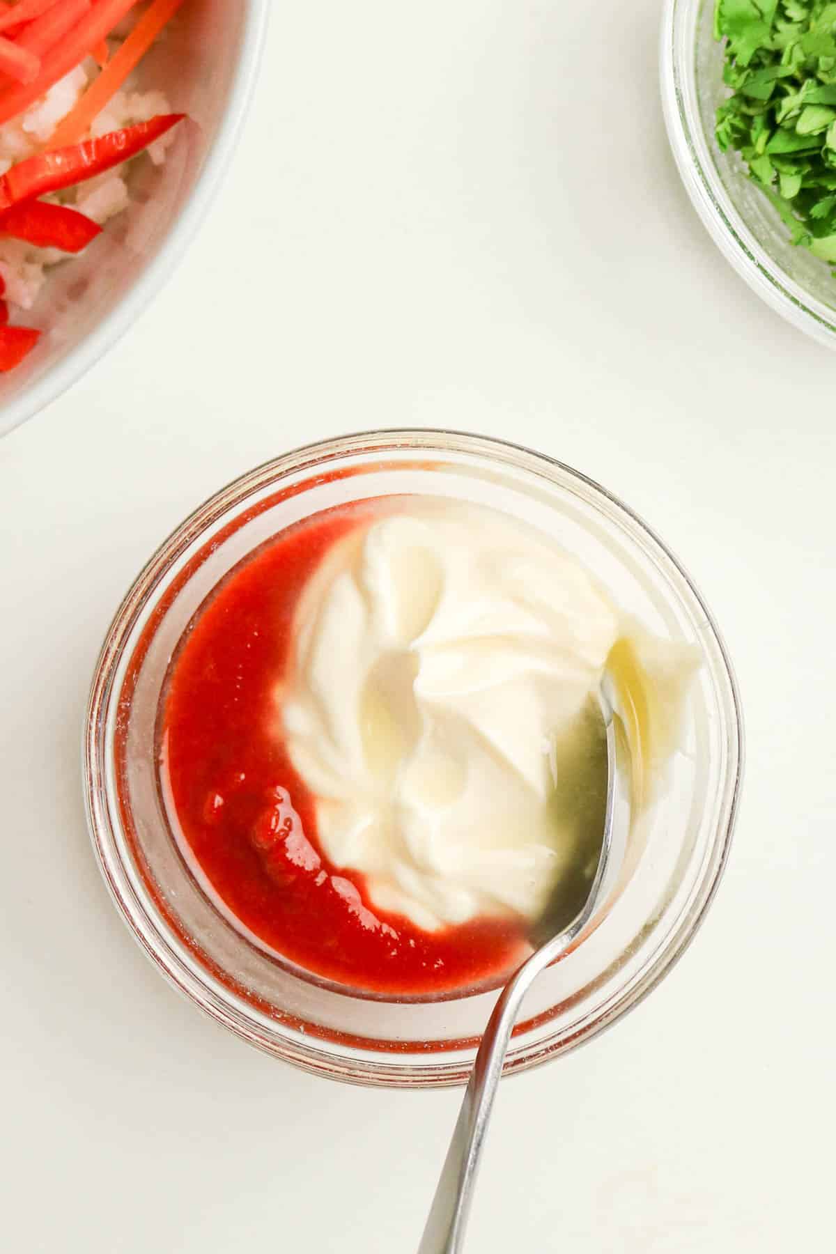 A spoon in a bowl of mayonnaise and ketchup, with cilantro and sliced vegetables nearby.