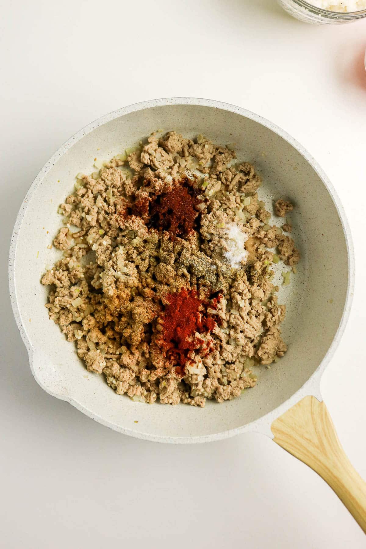 Ground meat cooking in a white skillet, topped with several different spices, on a white surface.