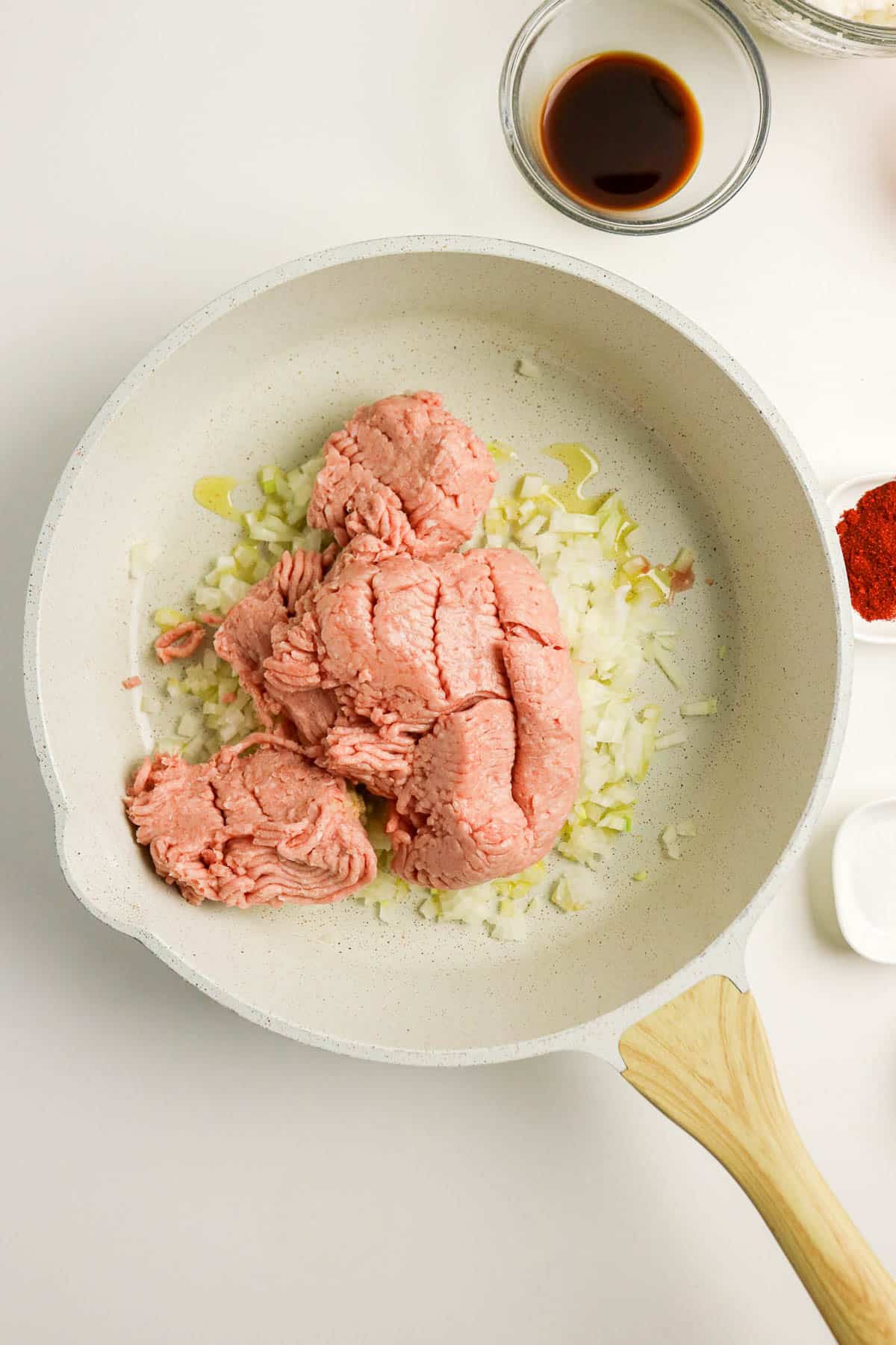 Raw ground turkey and chopped onions in a white skillet, with seasonings and sauce in bowls nearby.