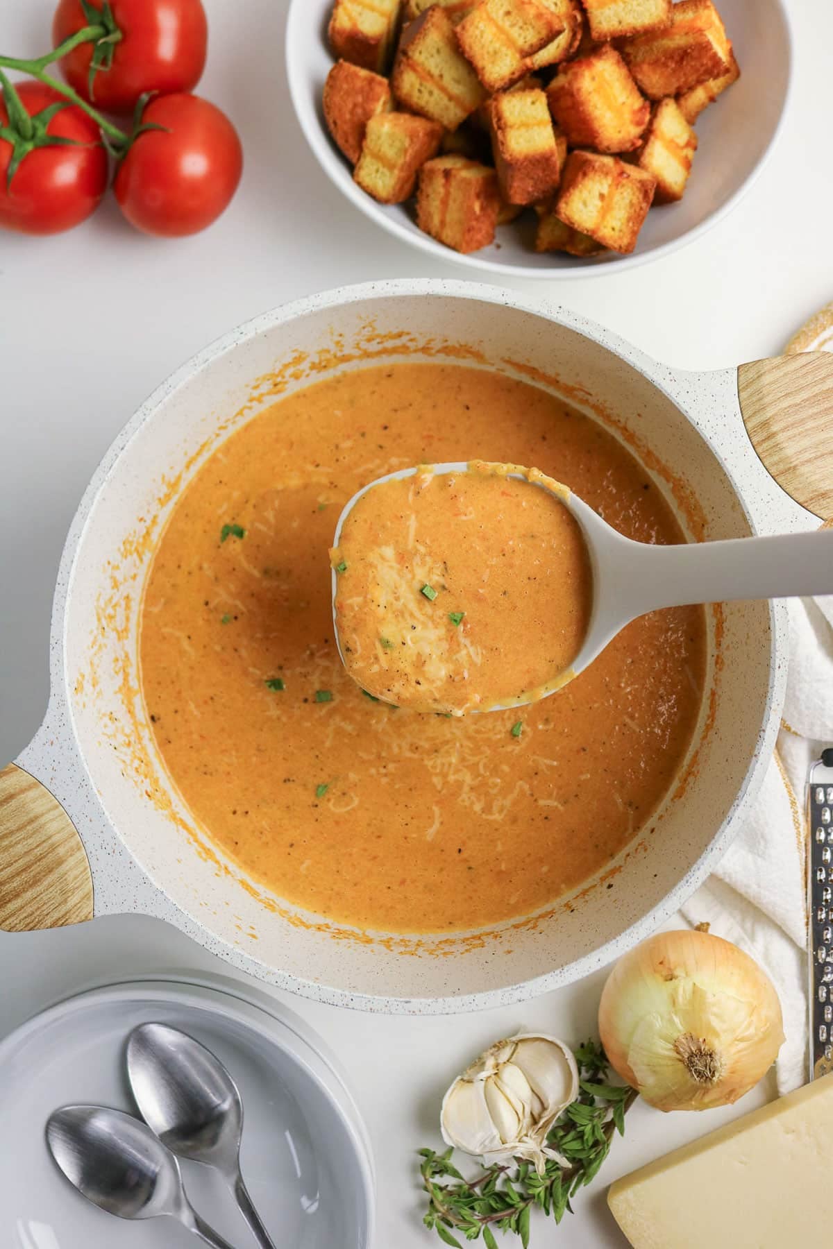 Ladle of creamy tomato soup in pot, with croutons, tomatoes, and soup ingredients on a white table.