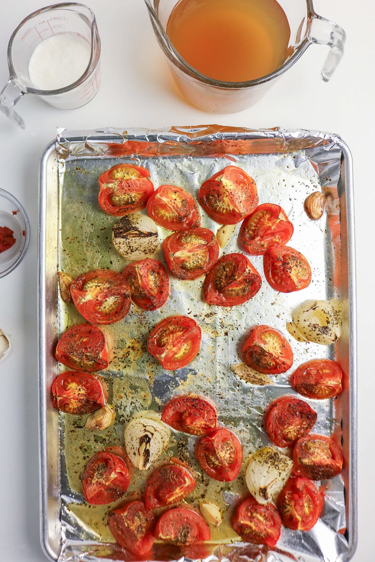 Halved tomatoes, onions, and garlic roasted on a foil-lined tray with spices; broth and seasonings nearby.